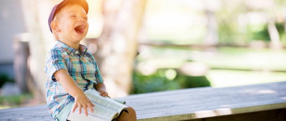 Ein kleiner Junge sitzt auf einer Bank mit einem Buch auf dem Schoß und lacht
